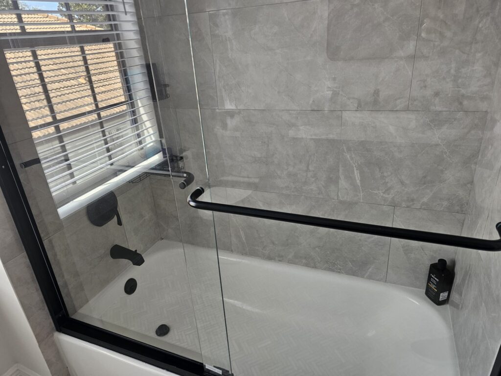 Glass-walled shower with gray marble tiles, black metal handles, and a bottle of shampoo on the tub ledge.